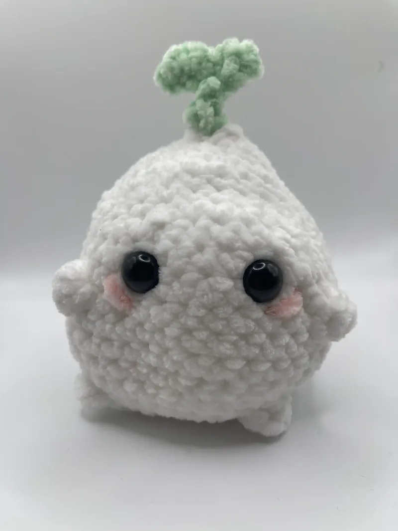 An ivory crochet onion with glossy black eyes, rosy cheeks, tiny feet, and a green sprout.