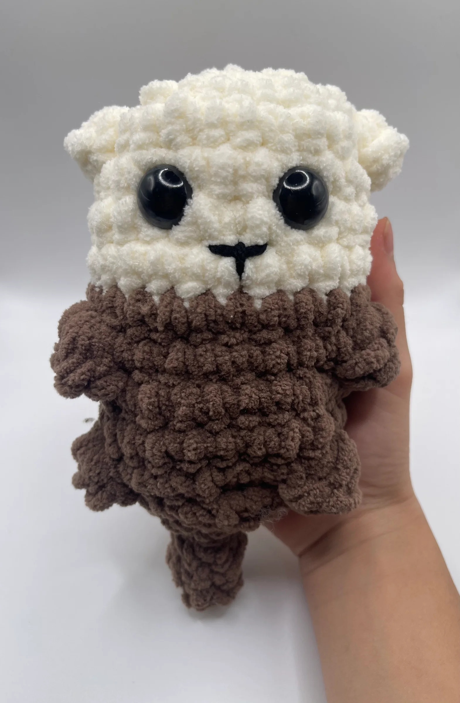 This upright animal plushie is crocheted with a cream-white head and small rounded ears, transitioning into a dark brown body with small side flippers and a textured tail at the base. Its face is detailed with glossy black safety eyes and a small, black T-shaped stitched nose and mouth.