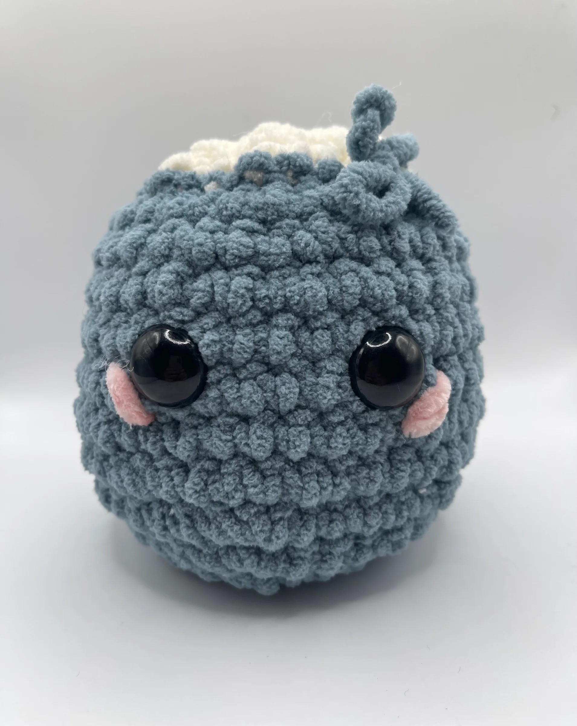 Handmade plushie crocheted from textured, dusty-blue chunky yarn and a white tufted top with a small bow. Its sweet face is detailed with large, glossy black eyes and soft pink blushing cheeks.